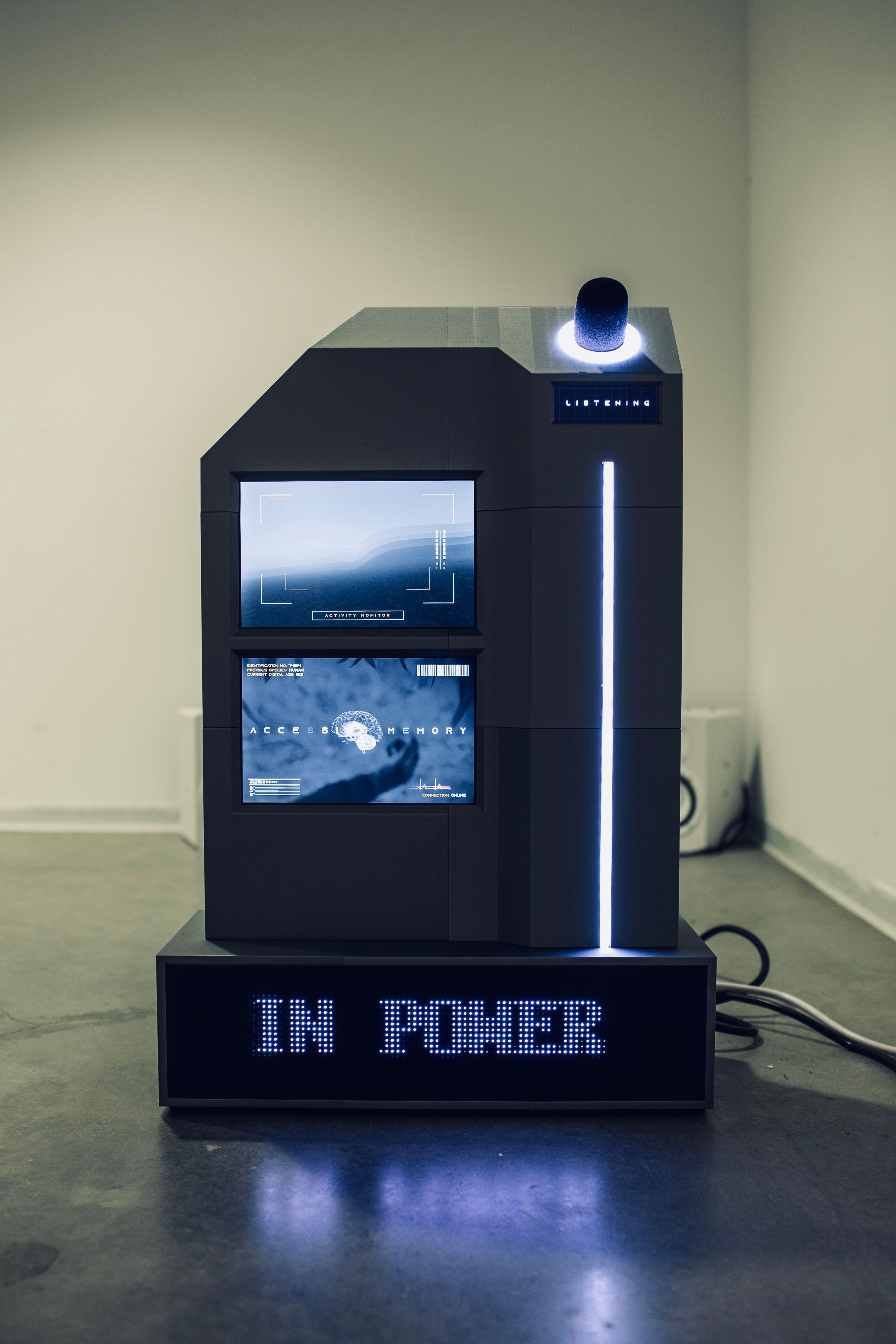 A geometric console sits on the floor of the gallery. It has two screens on the left side of the front panel, and on the right a column of light sits below a glowing knob. A the base of the console, a screen displays the phrase "In Power". 