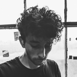 A black and white image of a young man with curly hair, wearing a black shirt, looking down at something. He is standing in front of a window that is divided into sections by thick window panes. There are some stickers on the glass. The man's face is most
