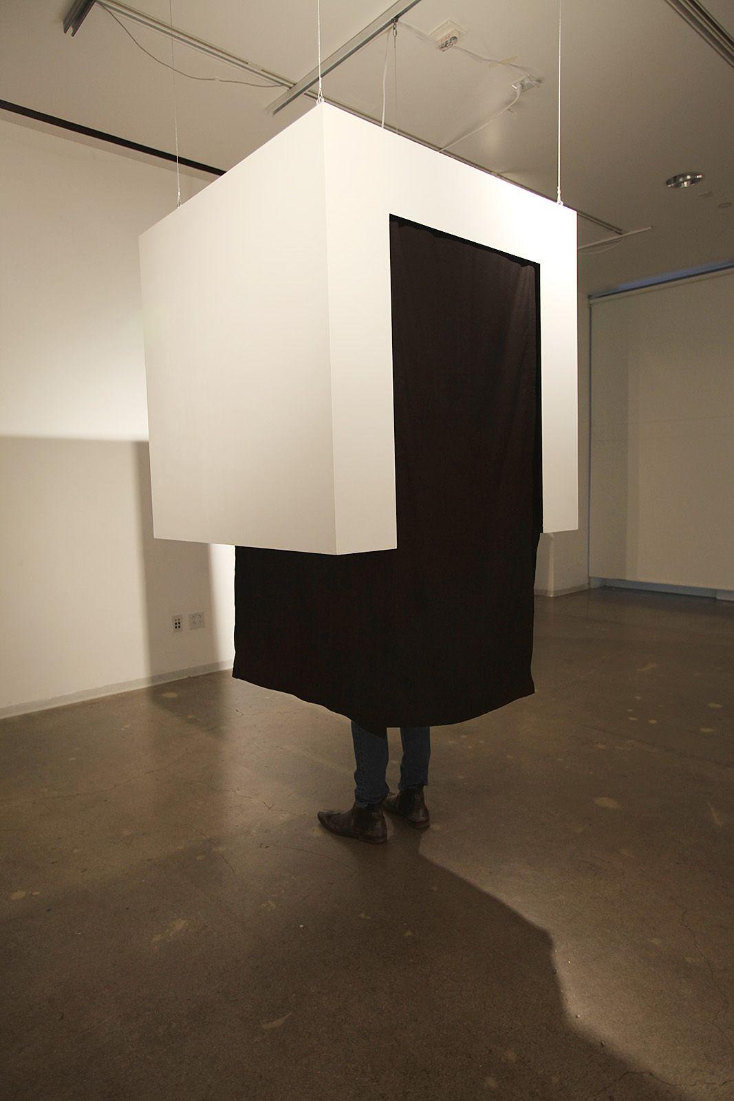 There is a white cube suspended from the air, with a black rectangle intersecting it. The black rectangle seems to be like a room inside the white cube. There are a pair of legs standing inside the white cube.