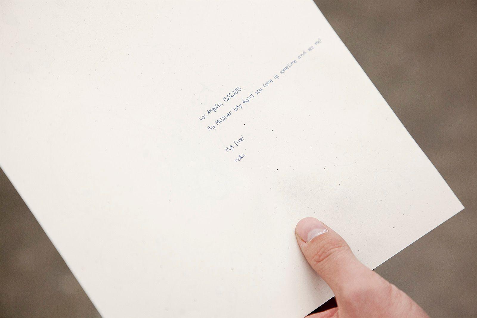 There is a paper held by a light skinned hand that has the text "Los Angeles, 13.02.2013". NEXT LINE: "Hey Matthias! Why don't you come up sometime and see me?" NEXT LINE: "High five!" NEXT LINE: "moka". The text is blue, small, and in a handwriting-like 