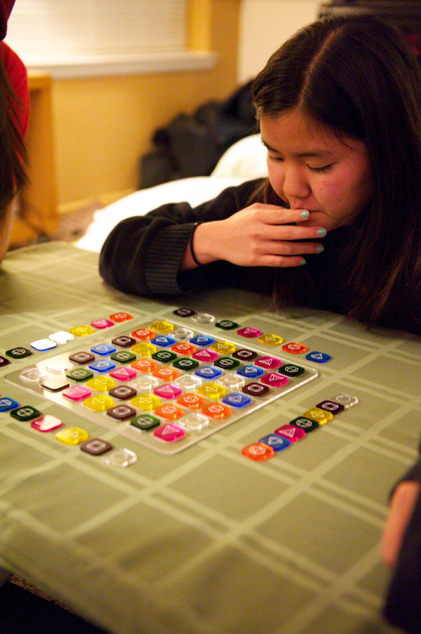 There is an Asian person looking down with their hand to their mouth that seems to be thinking. They are looking at a clear board filled with colorful square tokens arranged in a particular pattern.