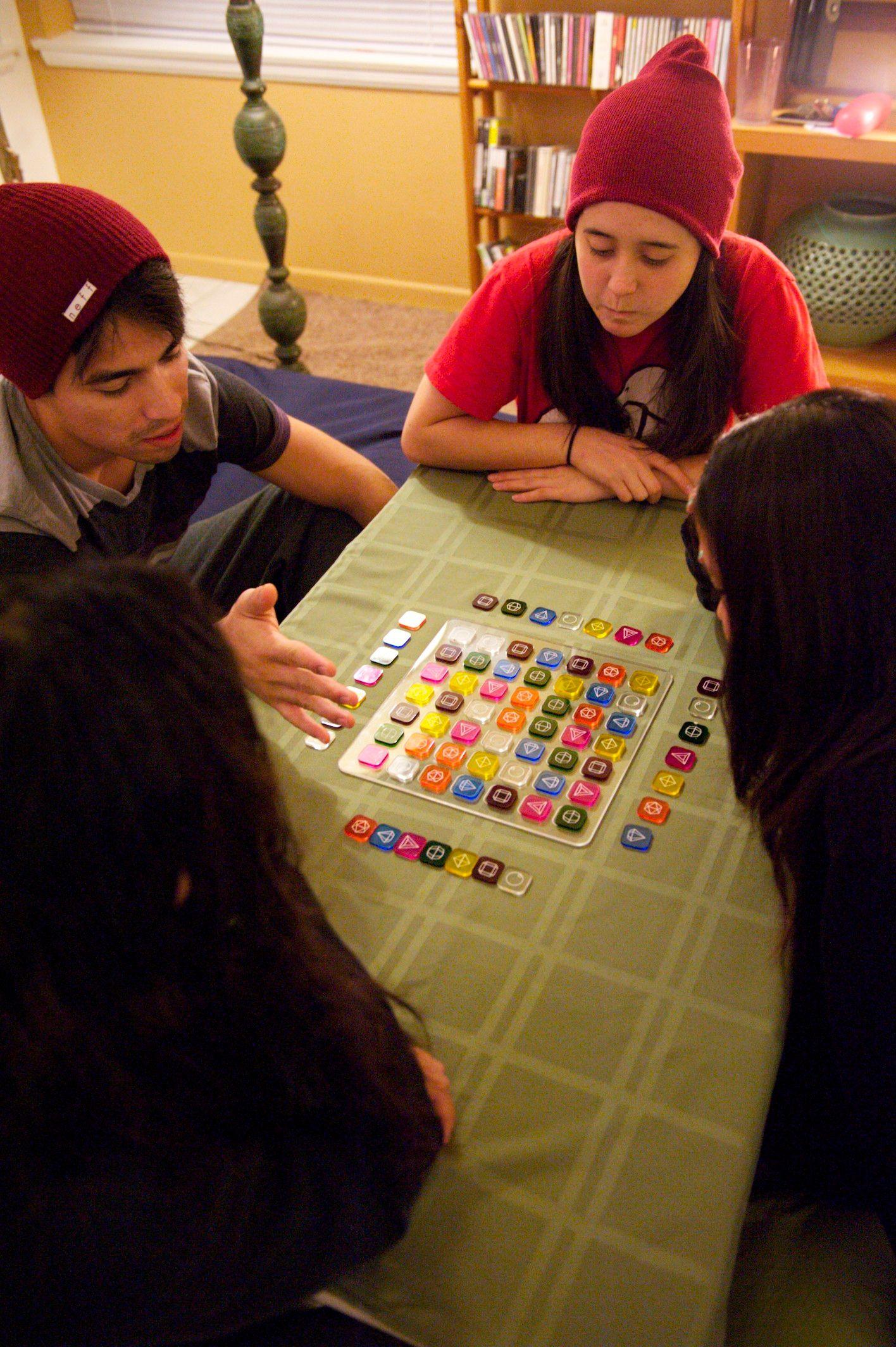 There are four people at a table with a clear square filled with colorful small square tokens in front of them. One person seems to be talking to the rest of the group.