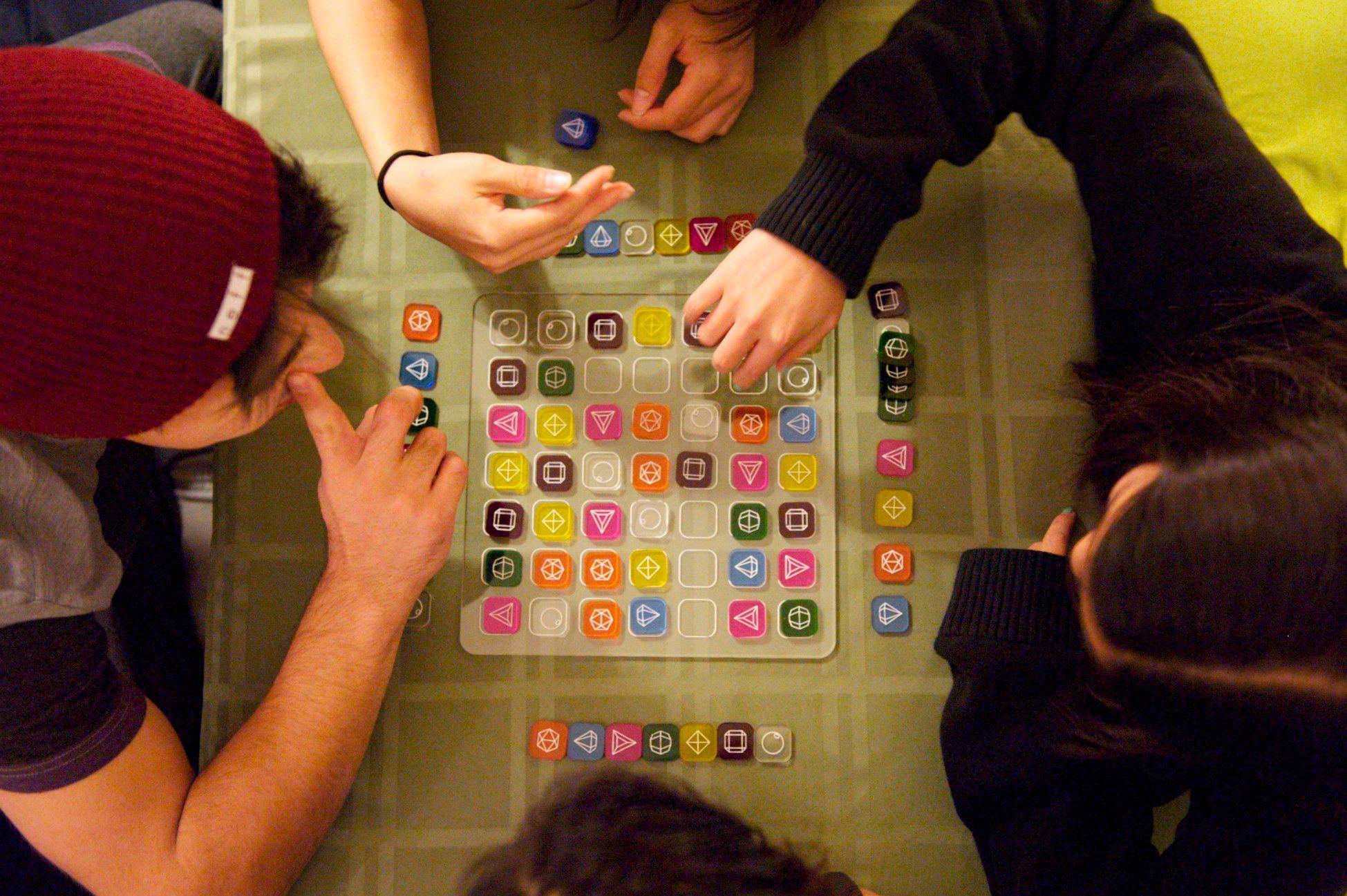 This is a birds eye view of four people handling colorful small square tiles on a larger clear square tile. 