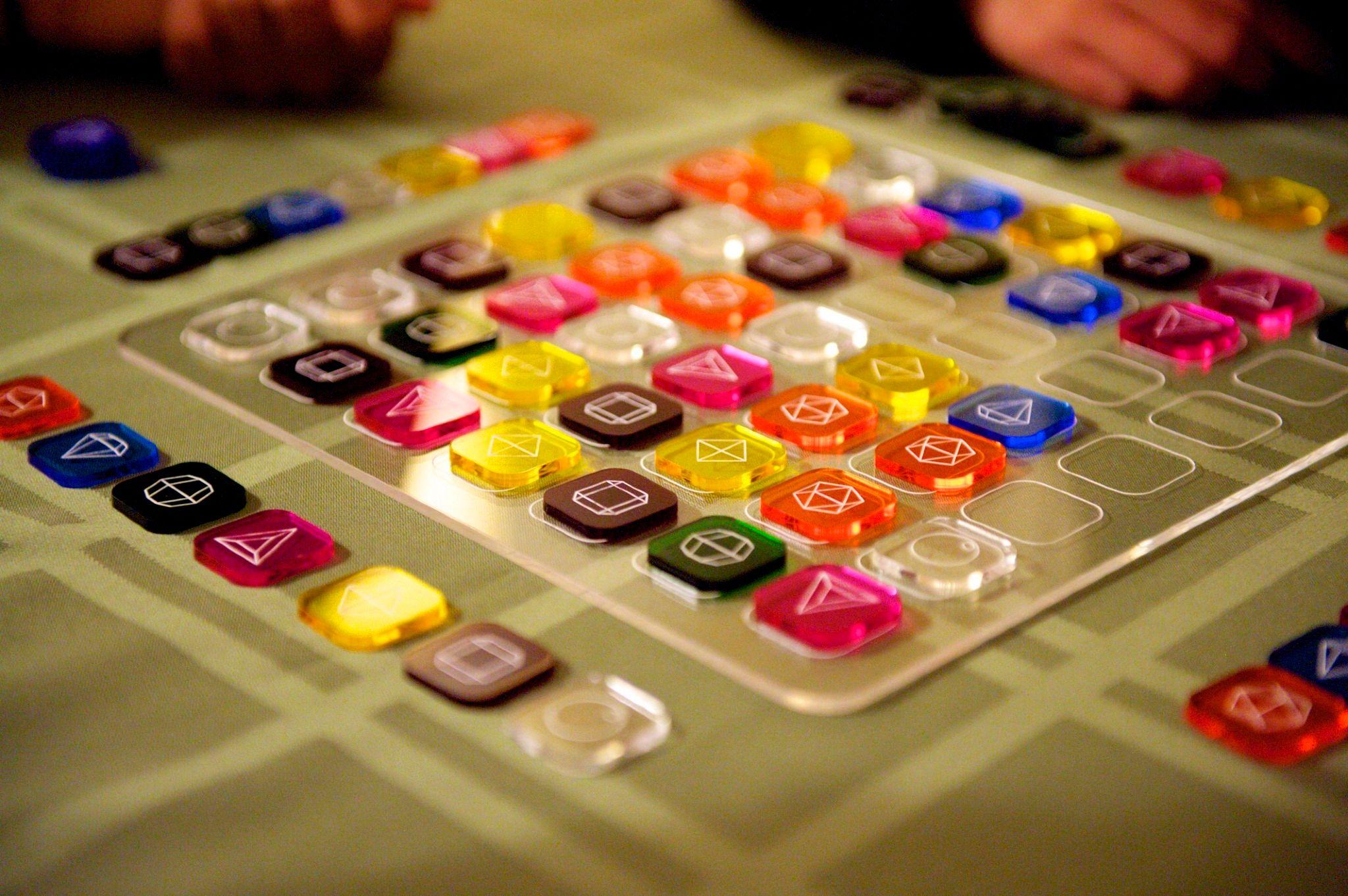 There are various square tokens on a clear white tile. Each of the tokens has a color and a shape unique to that color on it. There are green, brown, black, yellow, orange, clear, and dark pink tokens.