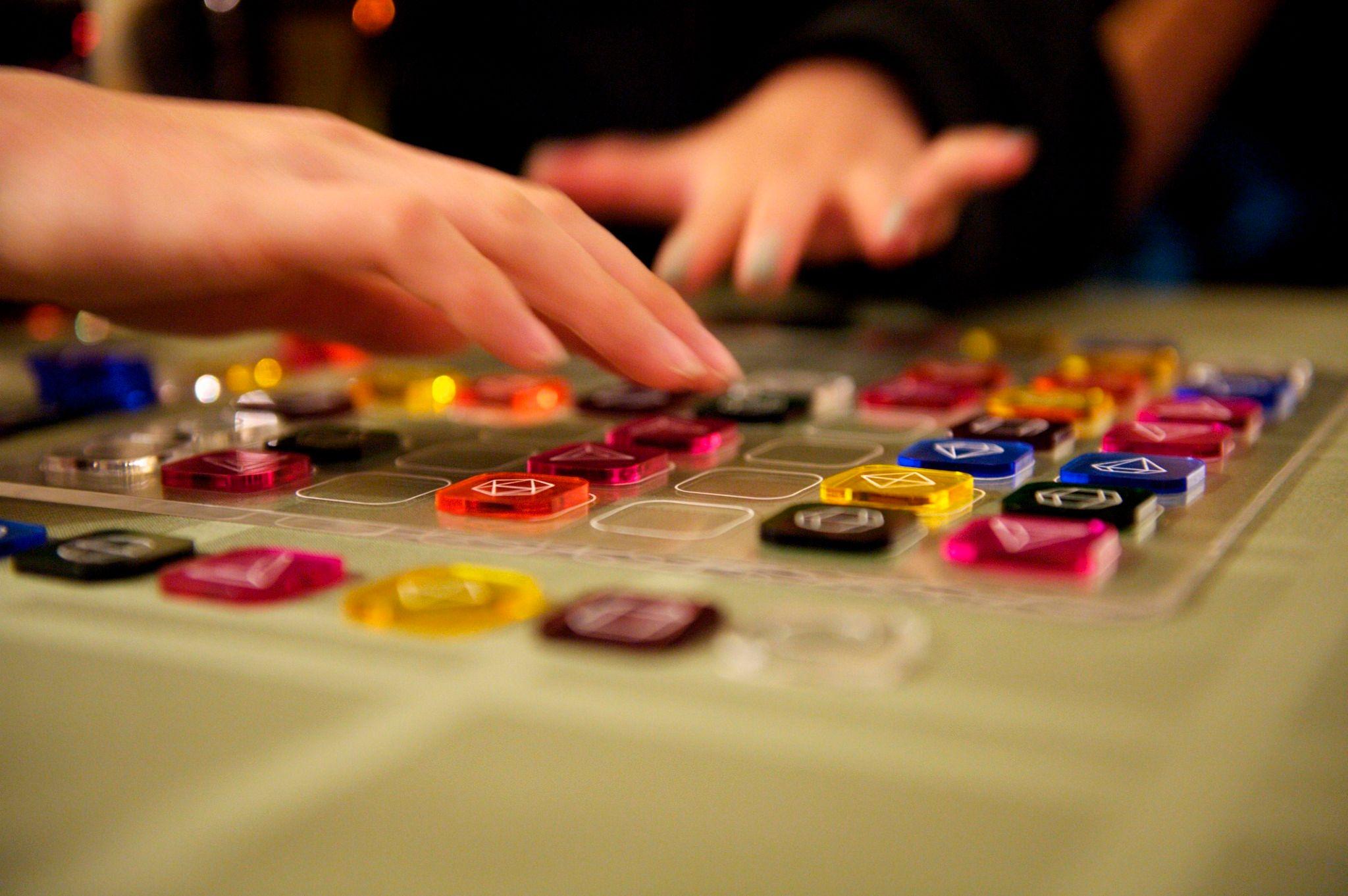 Two light skinned hands are reaching for colorful square tokens on a clear board.