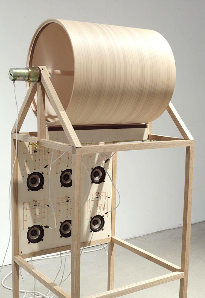 This is a light beige wooden machine with a cylinder on the top and six speakers on the bottom. Each speaker and the cylinder has one string attached to it. The machine is supported with thin beige wooden sticks.