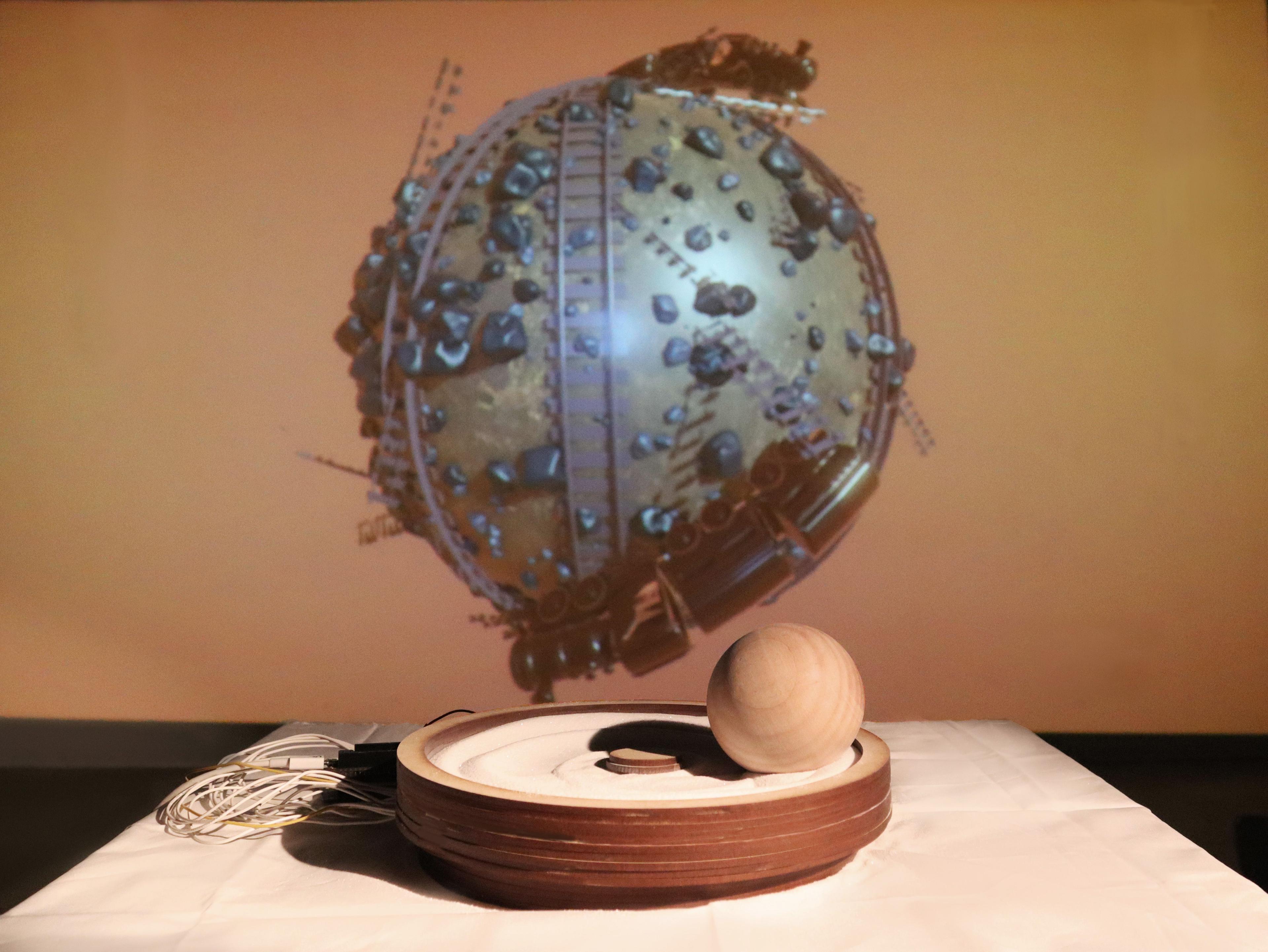 A projection screen with a 3d model of a small round planet. In the foreground there is a wooden vessel filled with sand. In the sand is a wooden ball.