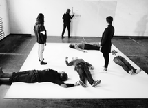 A black and white photo of performance lecture by the artist Julianna Johnston. Julianna is a white wall drawing a diagram of two points on an x and y axis. On the dark indoor floor there is a white square in which two participants are standing and anothe