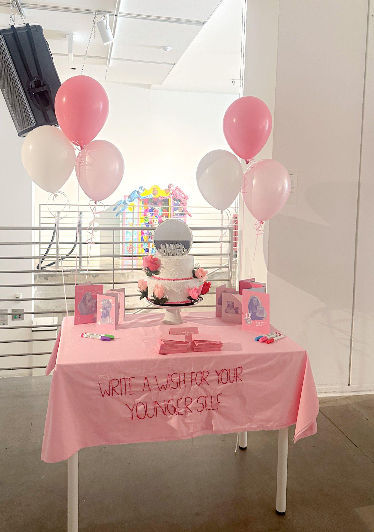 A table with a pink cover, on which "Write a wish for your younger self" is embroidered. On top of the table is a birthday cake, birtday cards, and multi-colored markers. Behind the table are two bundles of three balloons.