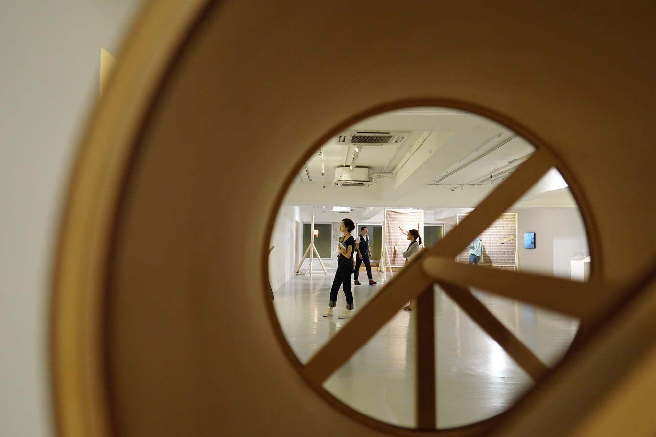 This is a photo taken inside a large cylinder so that only a portion of the actual room can be seen. The cylinder has wooden poles further covering some parts of the view. In the room are some people who seem to be observing things on the wall.