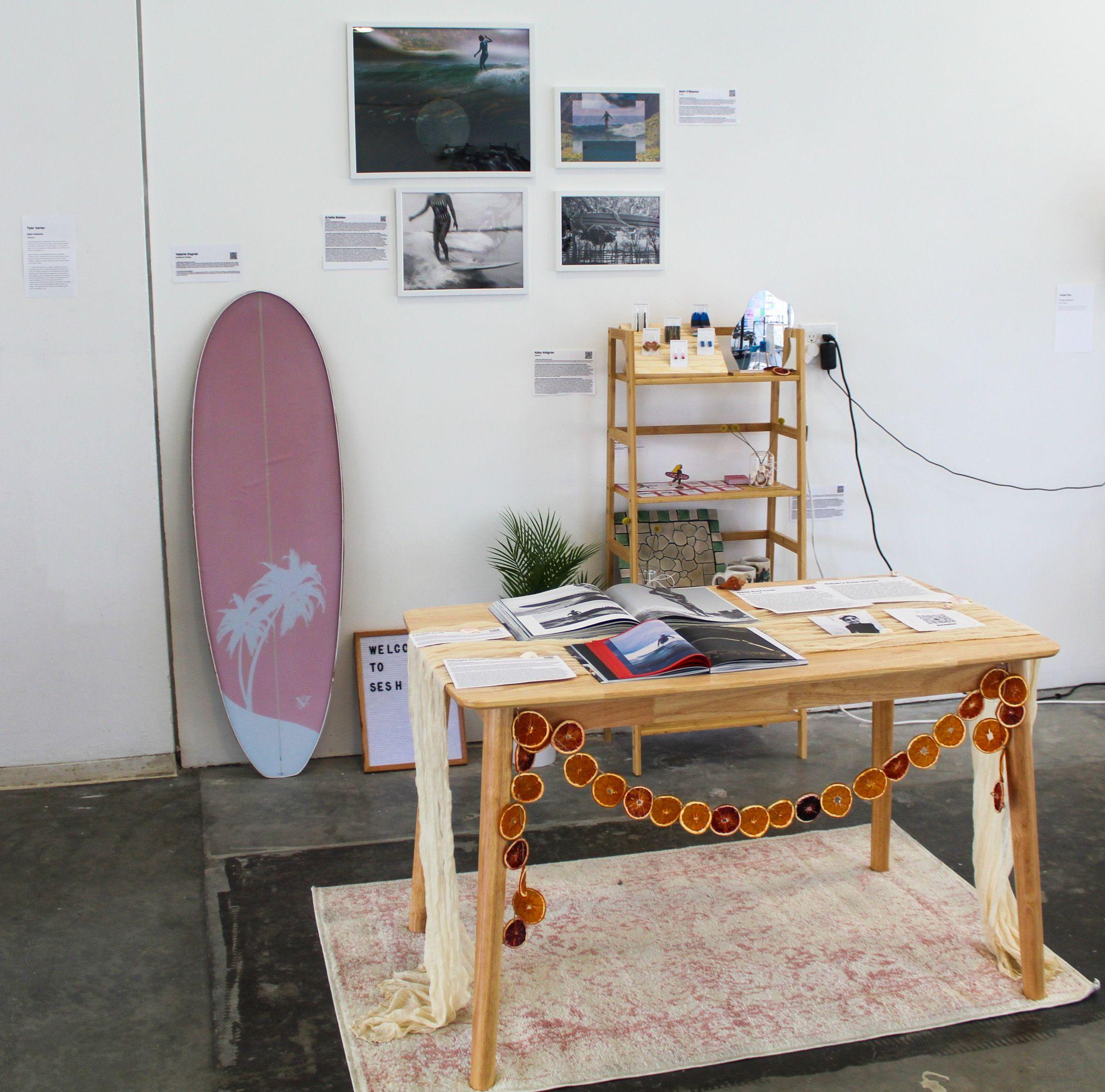 Surfing magazines lie open on a wooden table decorated with dried oranges strung to one another. Behind the table, a surfboard is propped against the wall next to a sign saying "Welcome to Sesh",  photographs of surfers, and a shelf with earrings and card