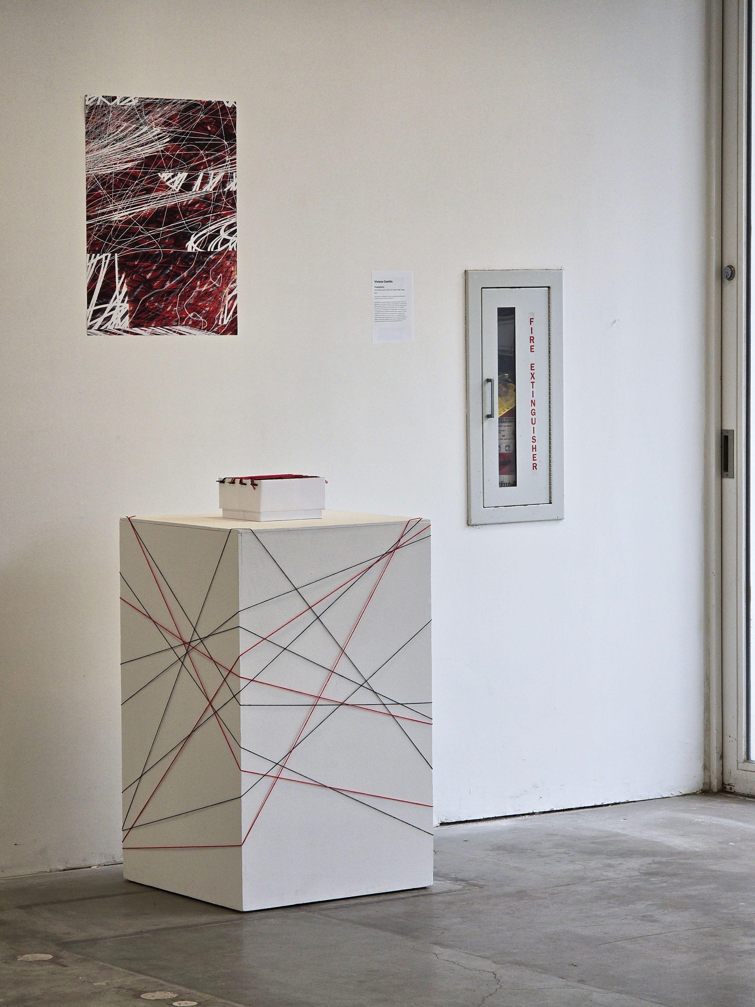 A podium wrapped in string with a similarly adorned box displayed on top of it. On the wall behind the podium, a poster with abstracted, web-like forms hangs. 