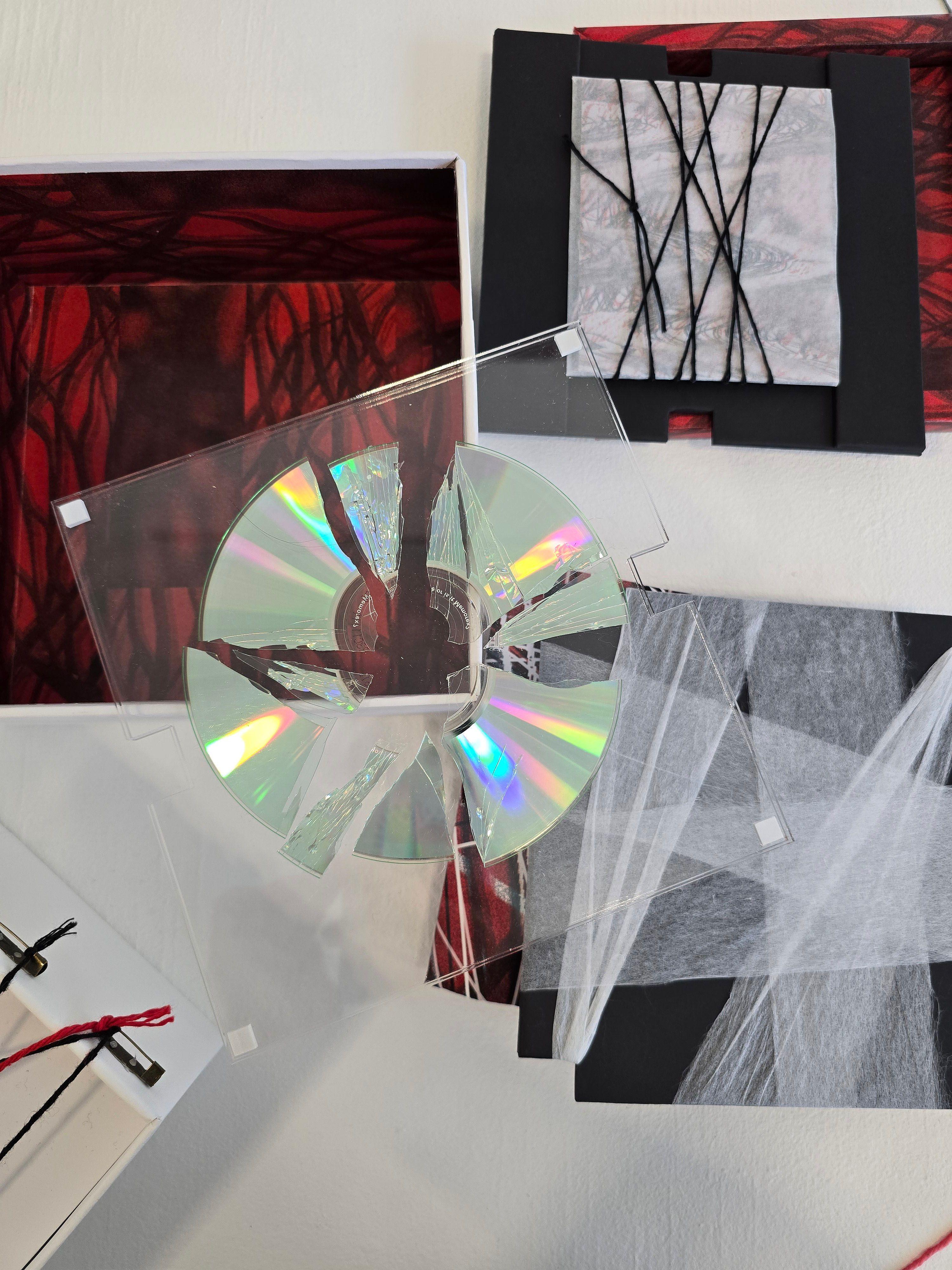 A CD is shattered into multiple shards, placed on top of a poster wrapped with strings and a zine. There is an open box with lined with a dark, swirling pattern, and the lid nearby.