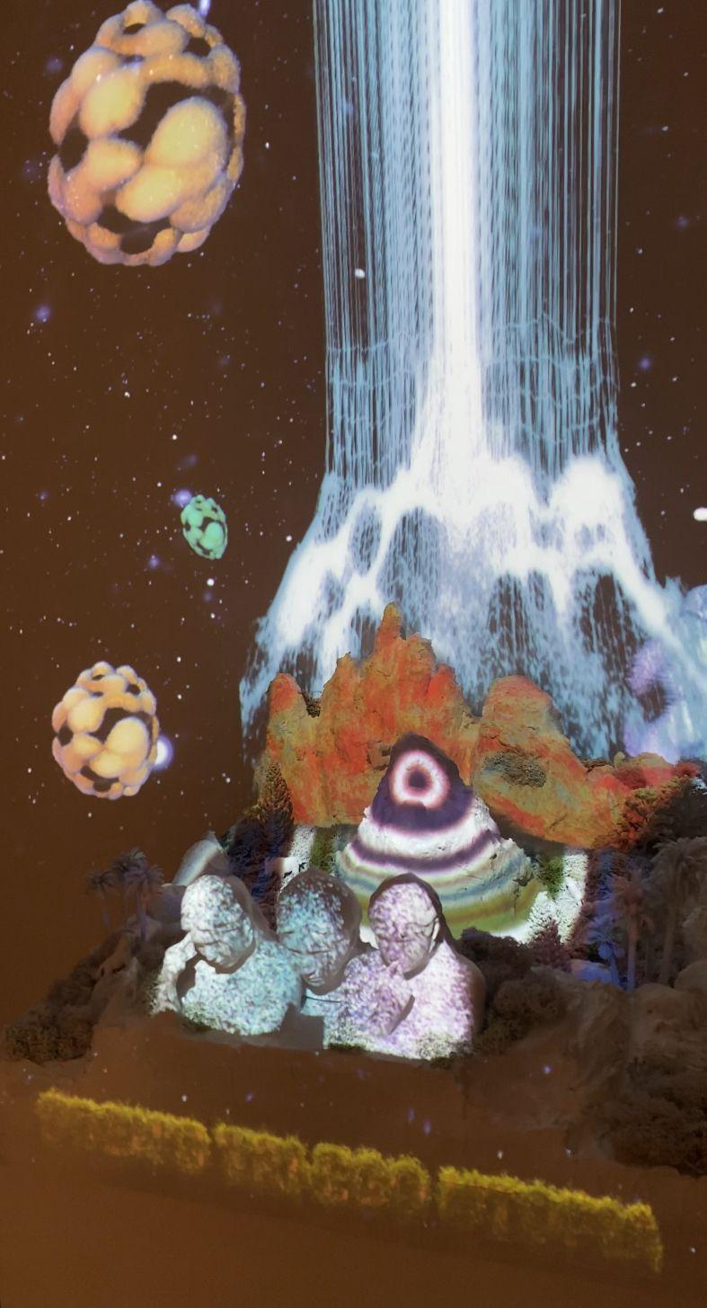 A beacon of vertical lines emerges from a lush, mountainous base, within which three human busts are placed. The scene is lit with a projection of layered colors appearing like sediment, and in the background, large, cellular masses of spheres float over 