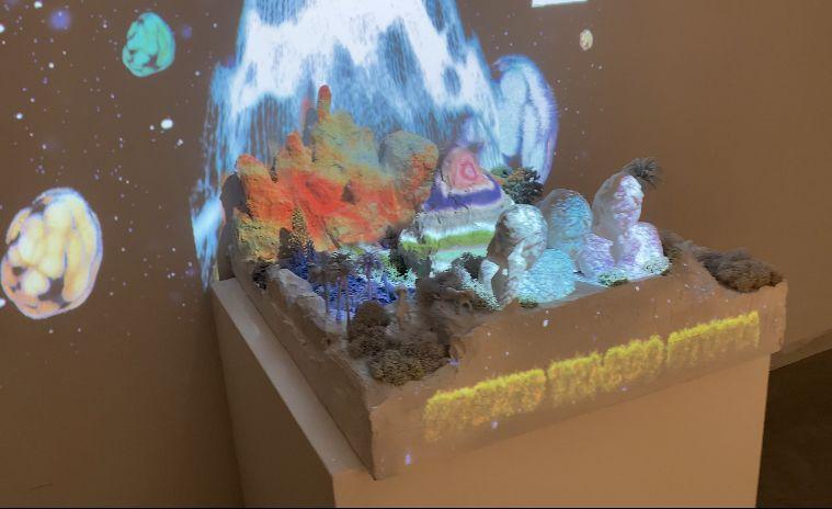 A close up of the beacon of vertical lines emerging from a lush, mountainous base, within which three human busts are placed. The scene is lit with a projection of layered colors appearing like sediment, and in the background, large, cellular masses of sp