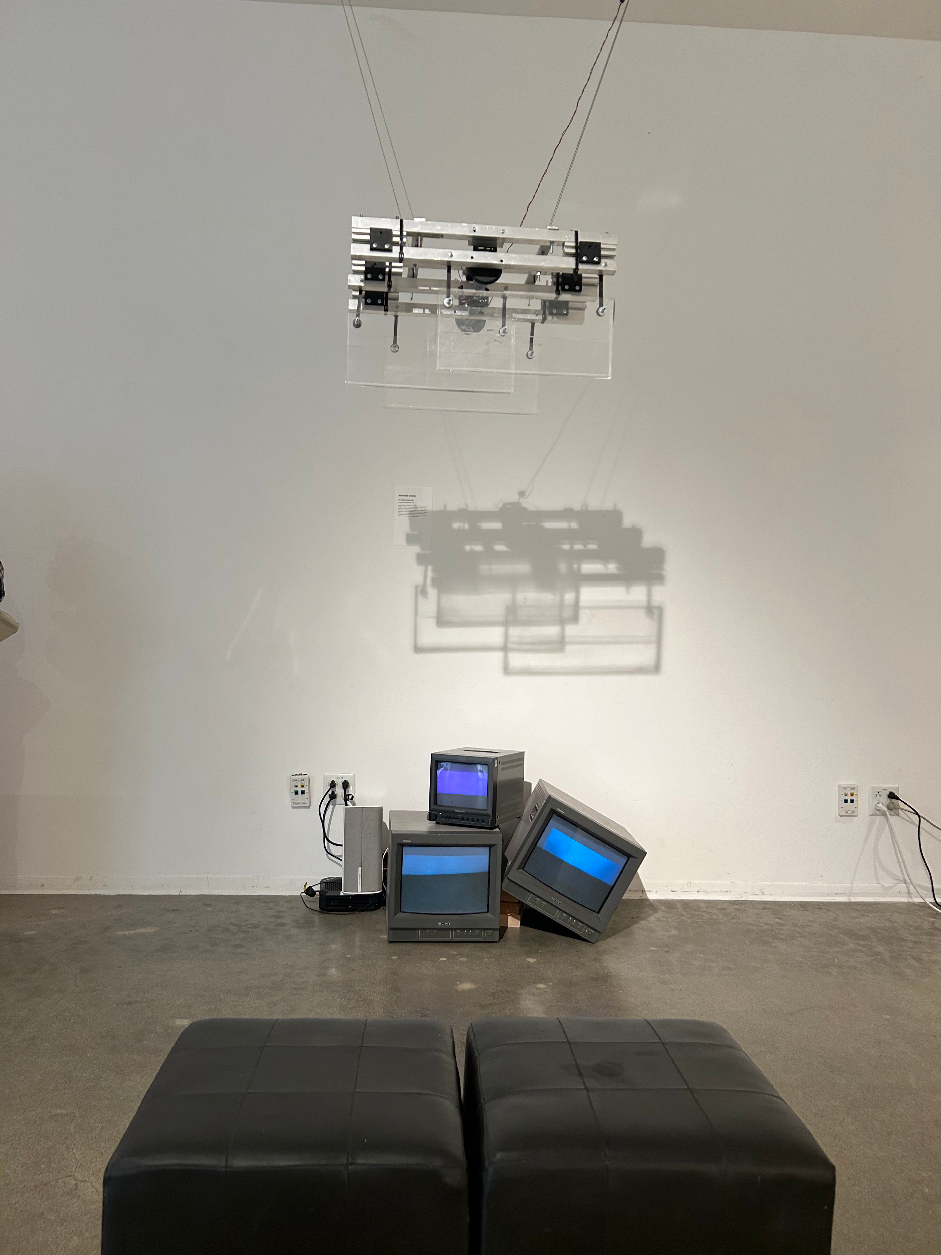 On the floor of the gallery, three CRT TV's sit, one leaning against the other, and another placed on top. Above the CRT TV's, a metal contraption with three moving panels of plexi-glass is suspended from the ceiling.