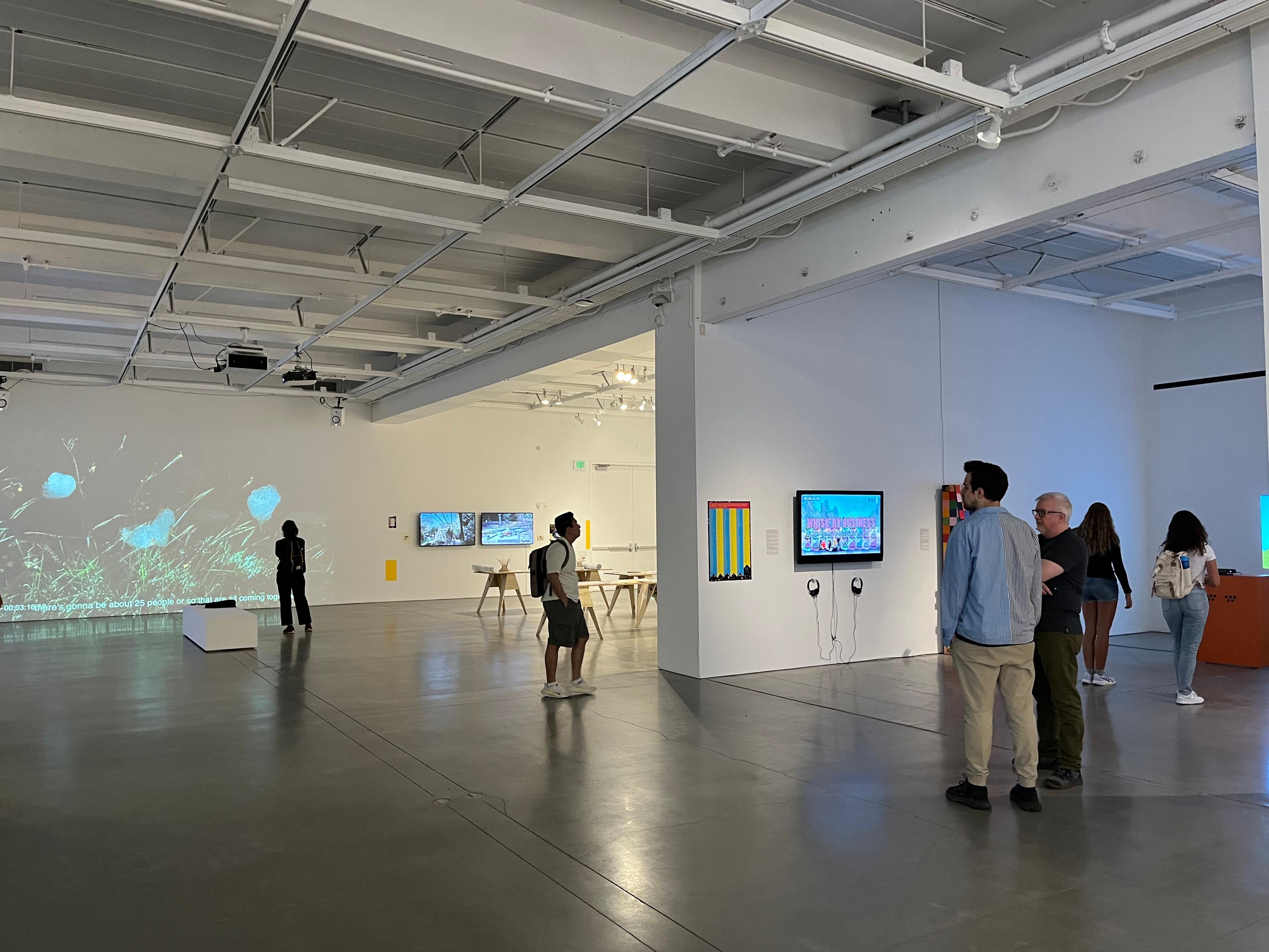 Visitors walk around the open exhibition space. In the background, a projection displays bundles of sporadically arranged lines. In the center of the image, a screen is mounted on the wall with two pairs of headphones hung beneath it.