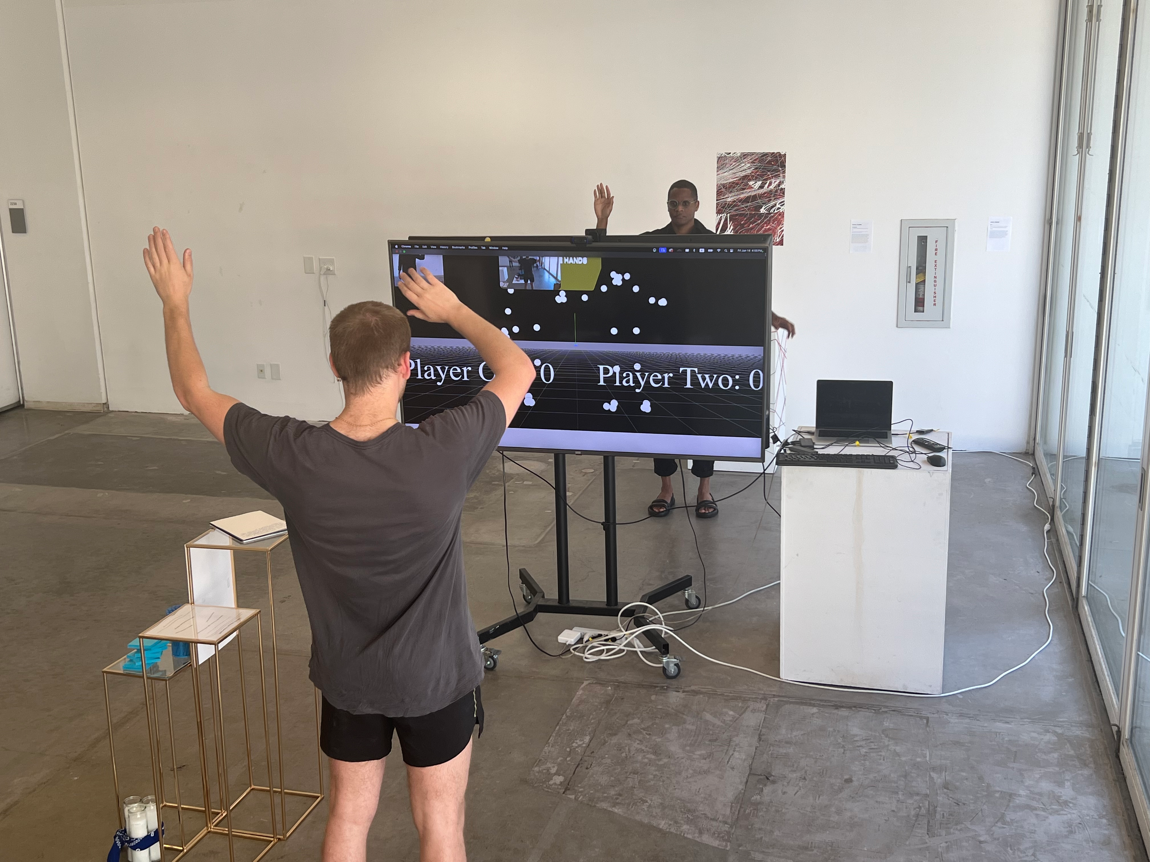 On opposite sides of two central screens, two individuals wave their hands around to grab spherical objects displayed on the screen. Movements are tracked by a camera in front of both participants, then translated into the game.