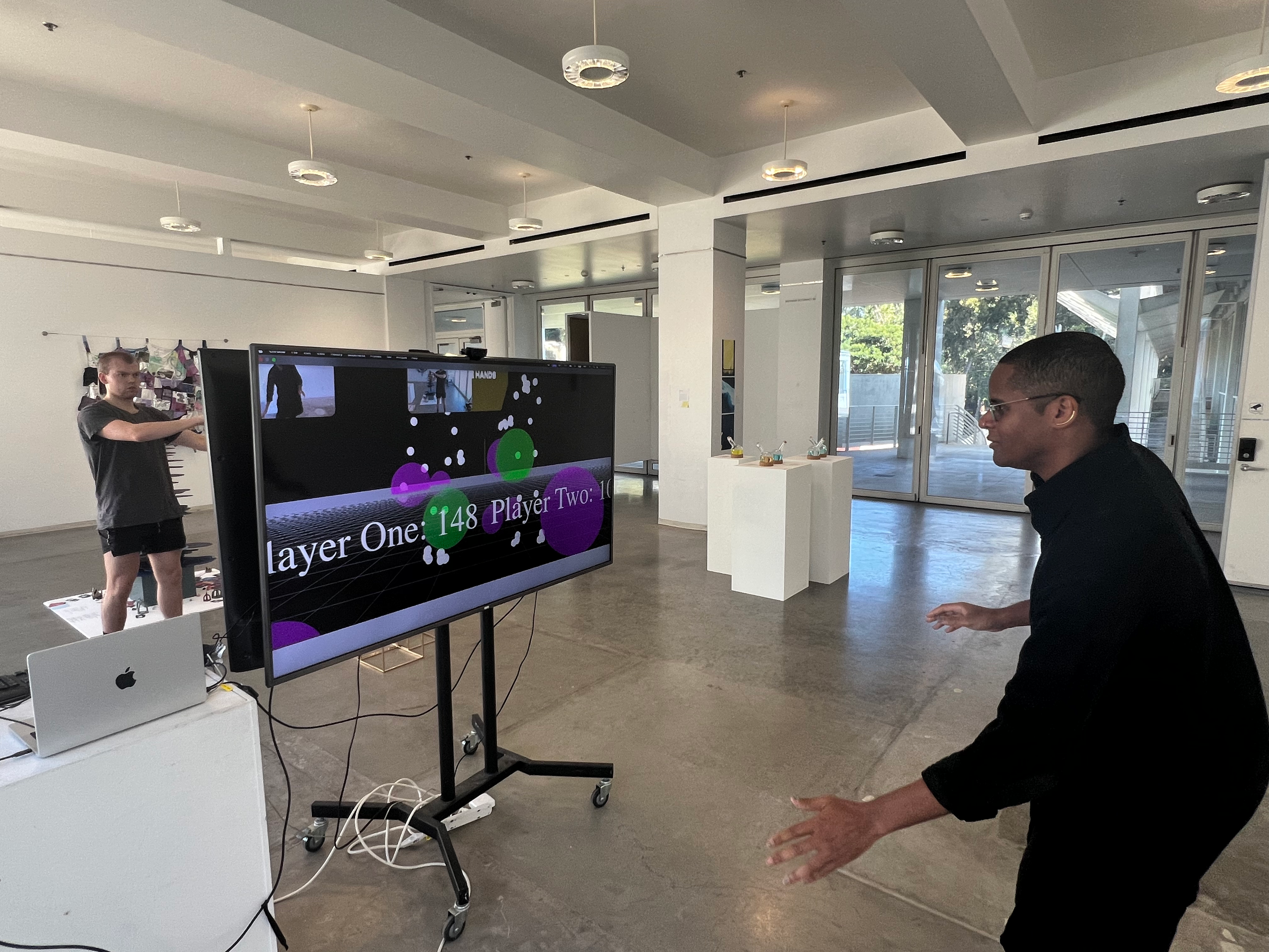 On opposite sides of two central screens, two individuals wave their hands around to grab spherical objects displayed on the screen. Movements are tracked by a camera in front of both participants, then translated into the game.