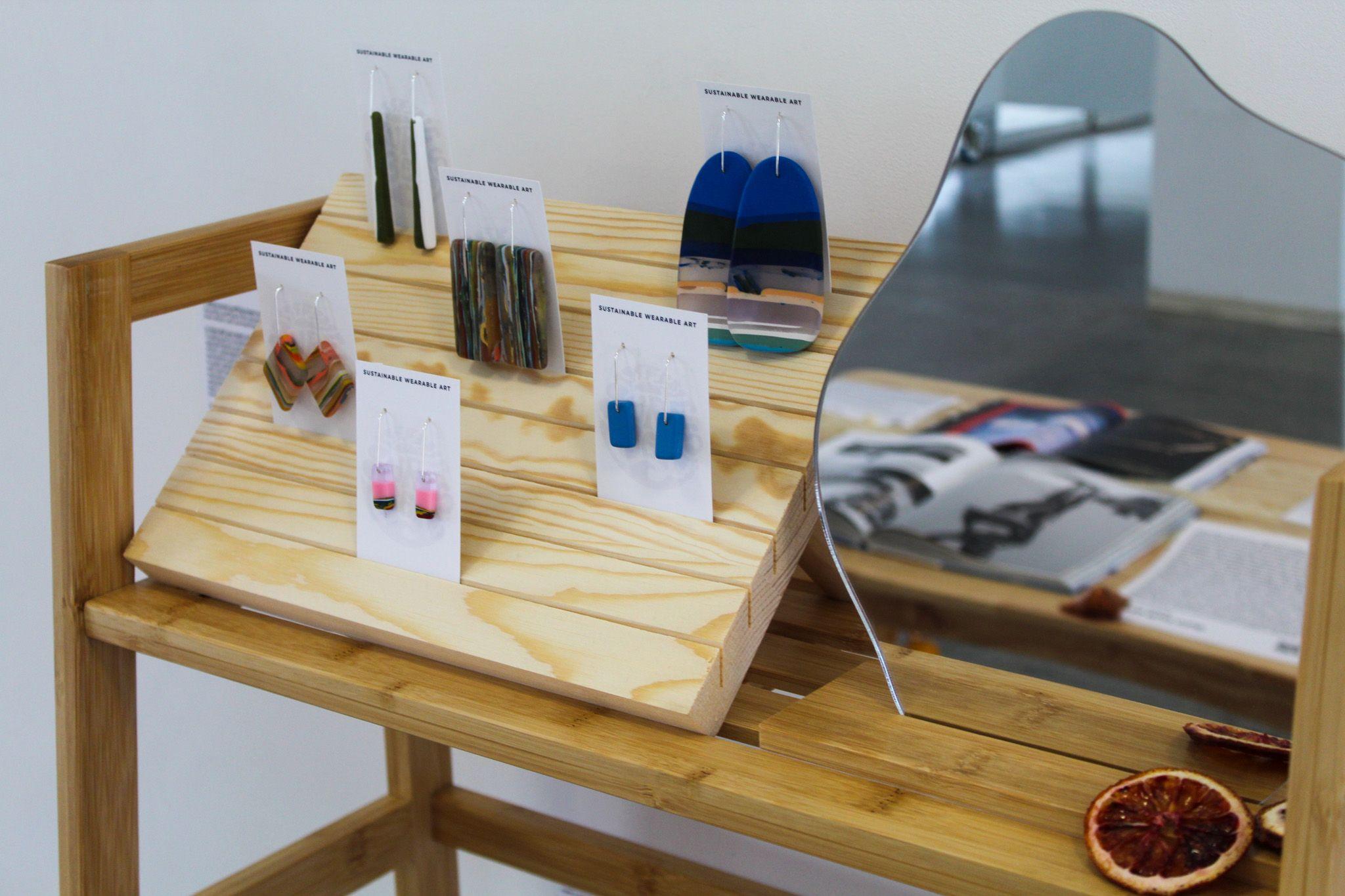 A close up image of the shelf shows gemstone earrings in a wooden display, next to an abstract, wavy mirror.