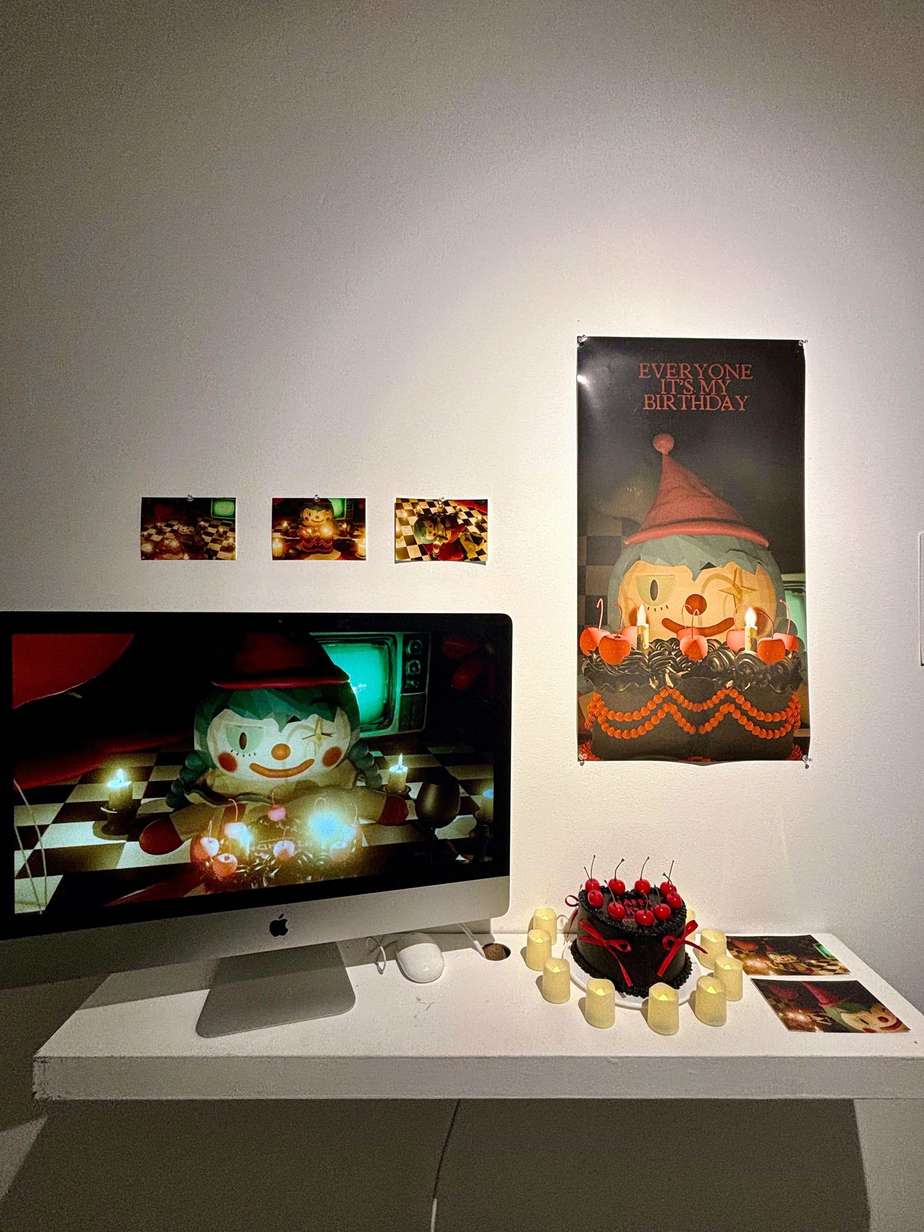 On a desk sits a monitor depicting a clown with a heart-shaped cake in front of it. The heart-shaped cake from the animation is placed next to the monitor, surrounded by candles. On the wall and the far right of the desk, photos of the animation are fixed