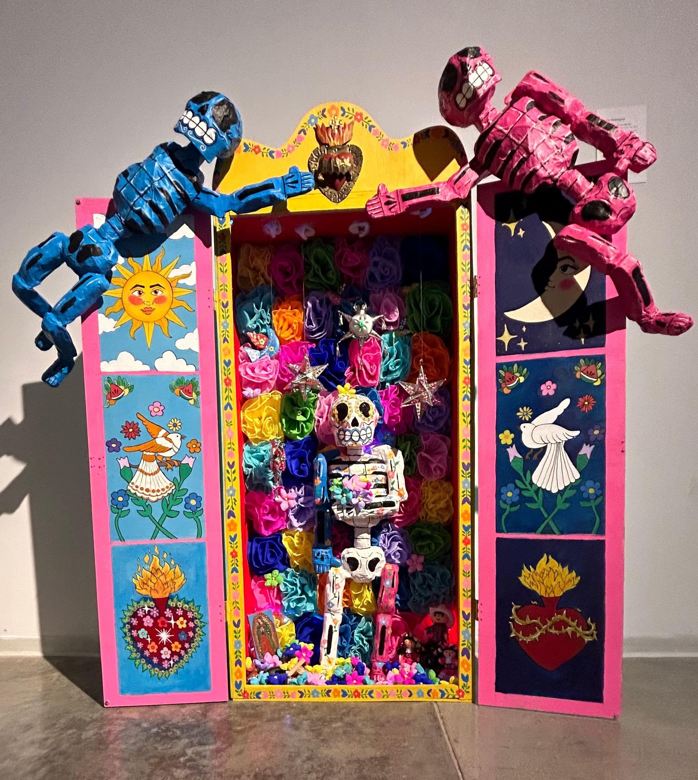 An open, brightly colored cabinet is lined with rows of multicolor flowers, within which a skeleton decorated with floral patterns stands. The interior of the cabinet doors are decorated with paintings of the sun and moon, doves, and a heart-shaped vessel