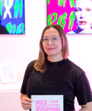 A white woman with shoulder-length brown hair, glasses, and a black shirt stands in a brightly lit space. They are holding a piece of paper with pink and black text. The background features vibrant, colorful digital artwork on display, with abstract shape