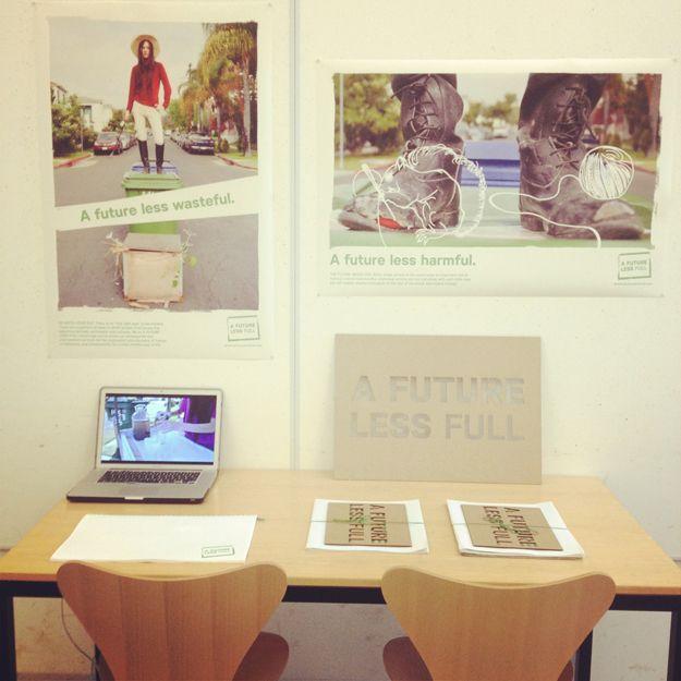 This is a straight-on photo of a desk against a wall. There are multiple materials all for the brand "A FUTURE LESS FULL" on the walls and on the table. There are two posters on the wall, and a laptop, two stacks of paper, and a small poster on the table.