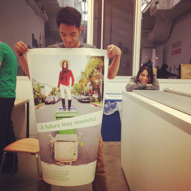There is a person holding a poster with a woman on top of a trashcan photographed, with the words "A future less wasteful." in green edited in. There is a woman watching and smiling.