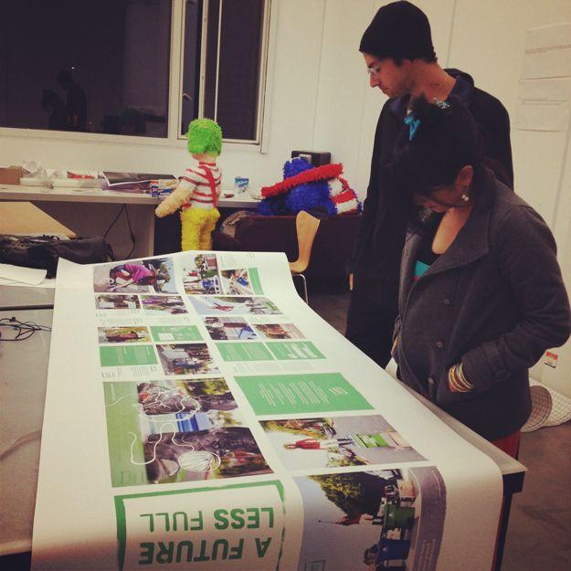 There are two people observing a large poster for "A FUTURE LESS FULL". The poster is larger than the table and has photos and green text.