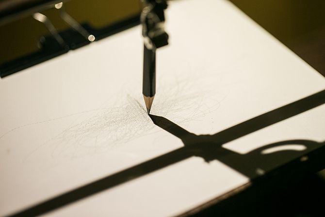 This is a picture of a pencil, shown by its shadow to be attached to a rod, drawing light, unintelligible lines on a white paper pad.