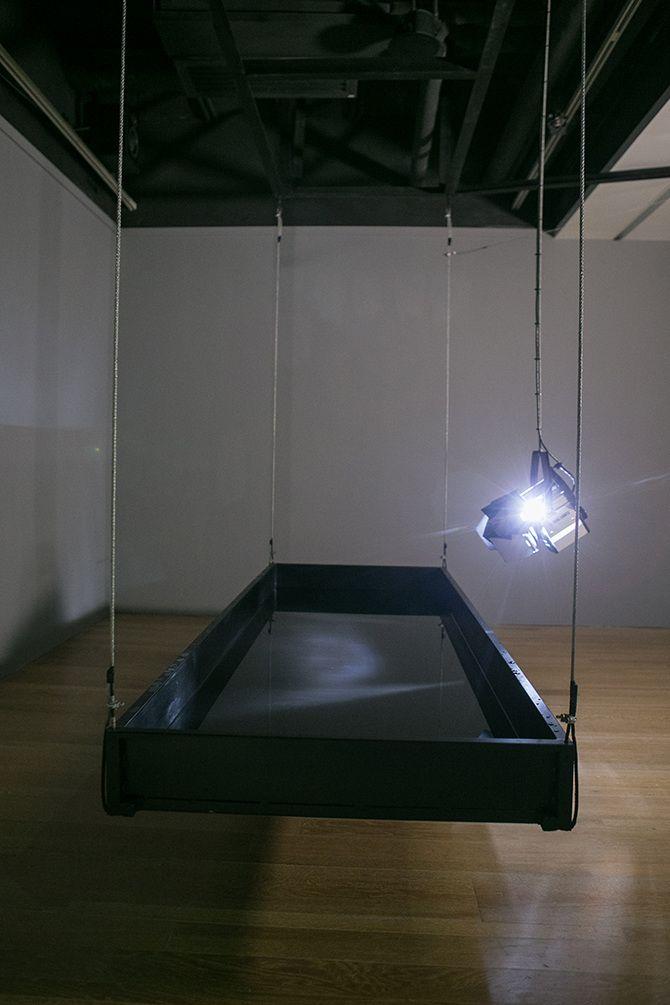 This is a photo of a stage light facing downward and a rectangular device suspended close to the ground and made of a black material.