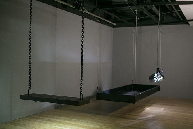 This is a diagonally angled photo of two main objects: a swing and a stage light. The swing is dark wood and has two chains to support it on either side. The stage light is angled downwards, facing the floor, and has a rectangular shaped box suspended in 