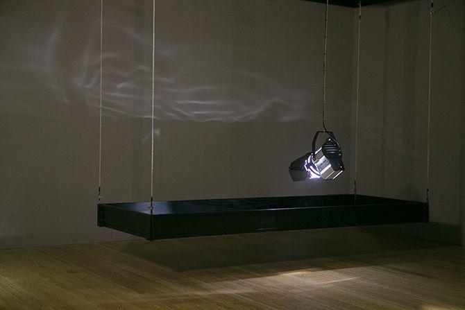 This is a photo of a stage light that is relatively close to the ground pointing down. The stage light has a rectangular box of black material suspended in the air around it. There are some abstract light reflections on the wall.
