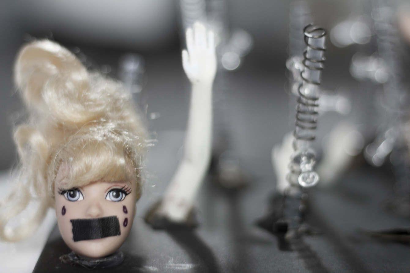 There is a Barbie doll head with black tape on its mouth and three black tears drawn on the cheeks. In the background is a portion of a doll arm and a small clear column with wire around it.