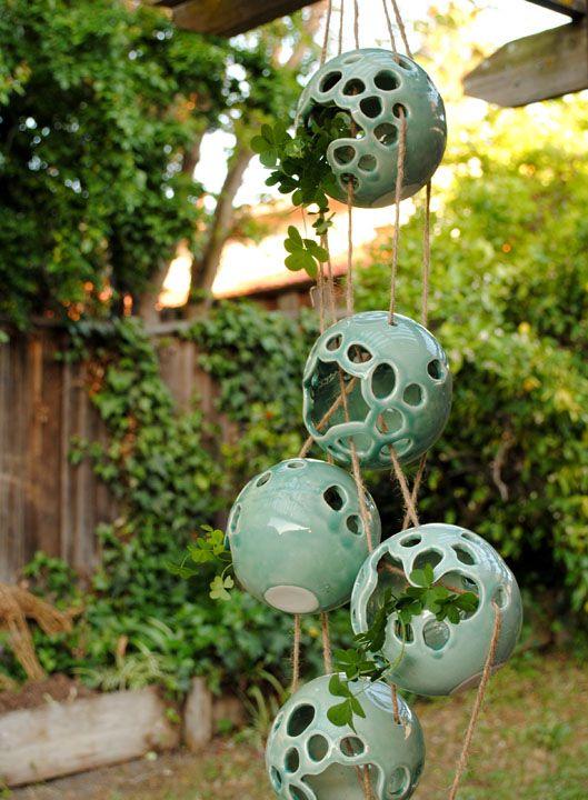 There are five green spherical ceramic objects with lots of holes inside them. There are clovers sprouting from them, and they are arranged to hang from the ceiling with twine. The photo was taken in a backyard like background on a sunny day.