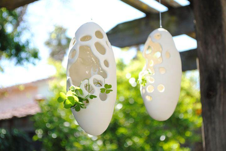 There are two white pods with lots of holes and one large hole in each hanging from a wooden structure. The photo was taken outside on a sunny day. There are clovers sprouting out of the objects.