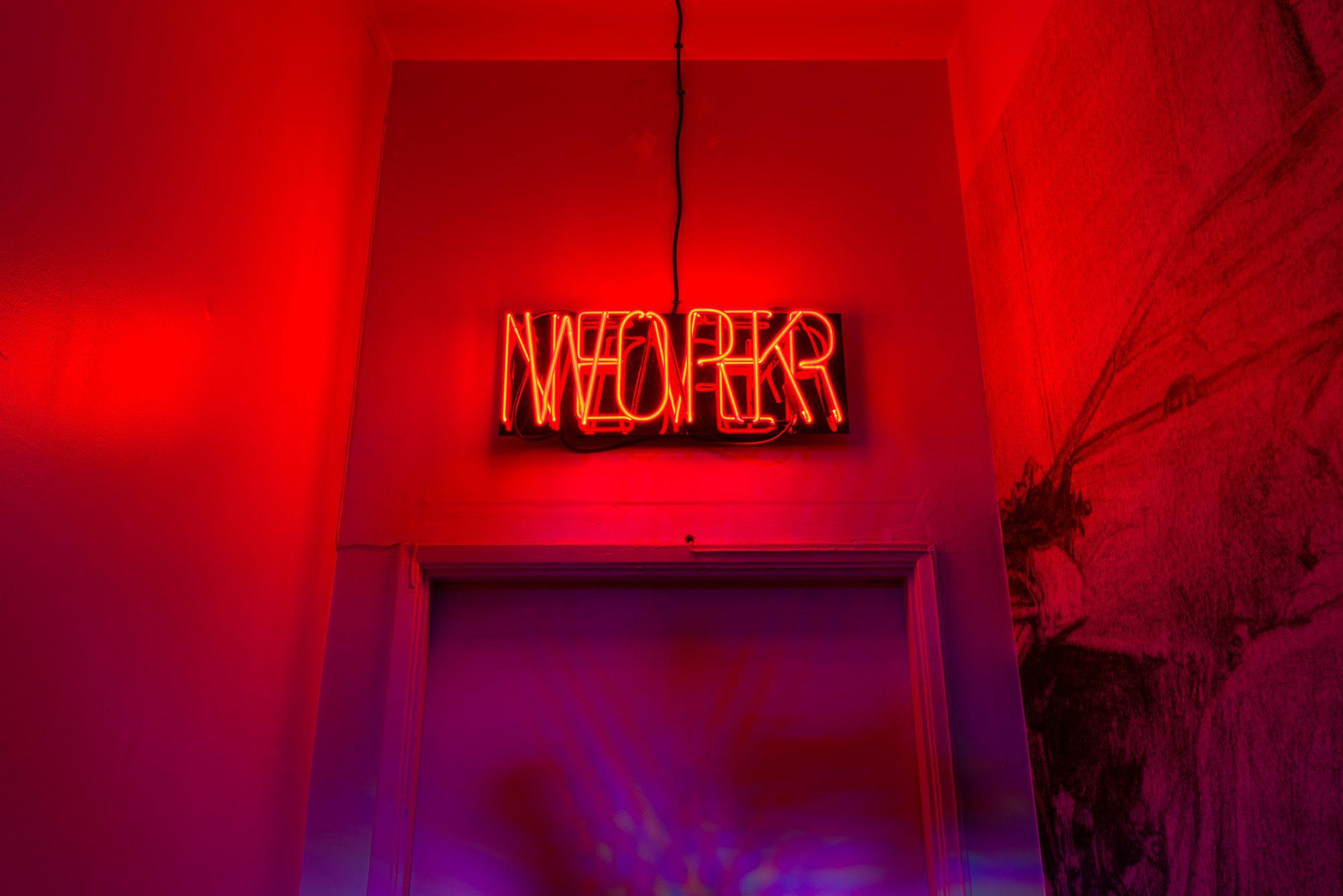 Overlapping words "NEVER WORK" in blood red neon, mounted above a door next to a large wall drawing.