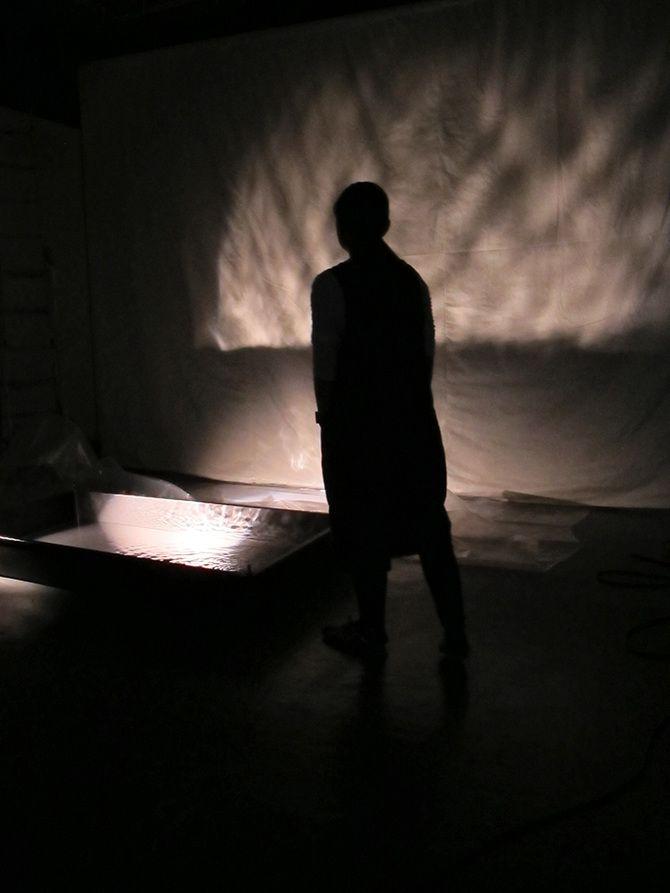 This is a photo of a person as the subject. There is a pan of what seems to be a liquid that is illuminated, and its reflection is illuminating the wall. The person is not lit, so only the shadow can be seen.