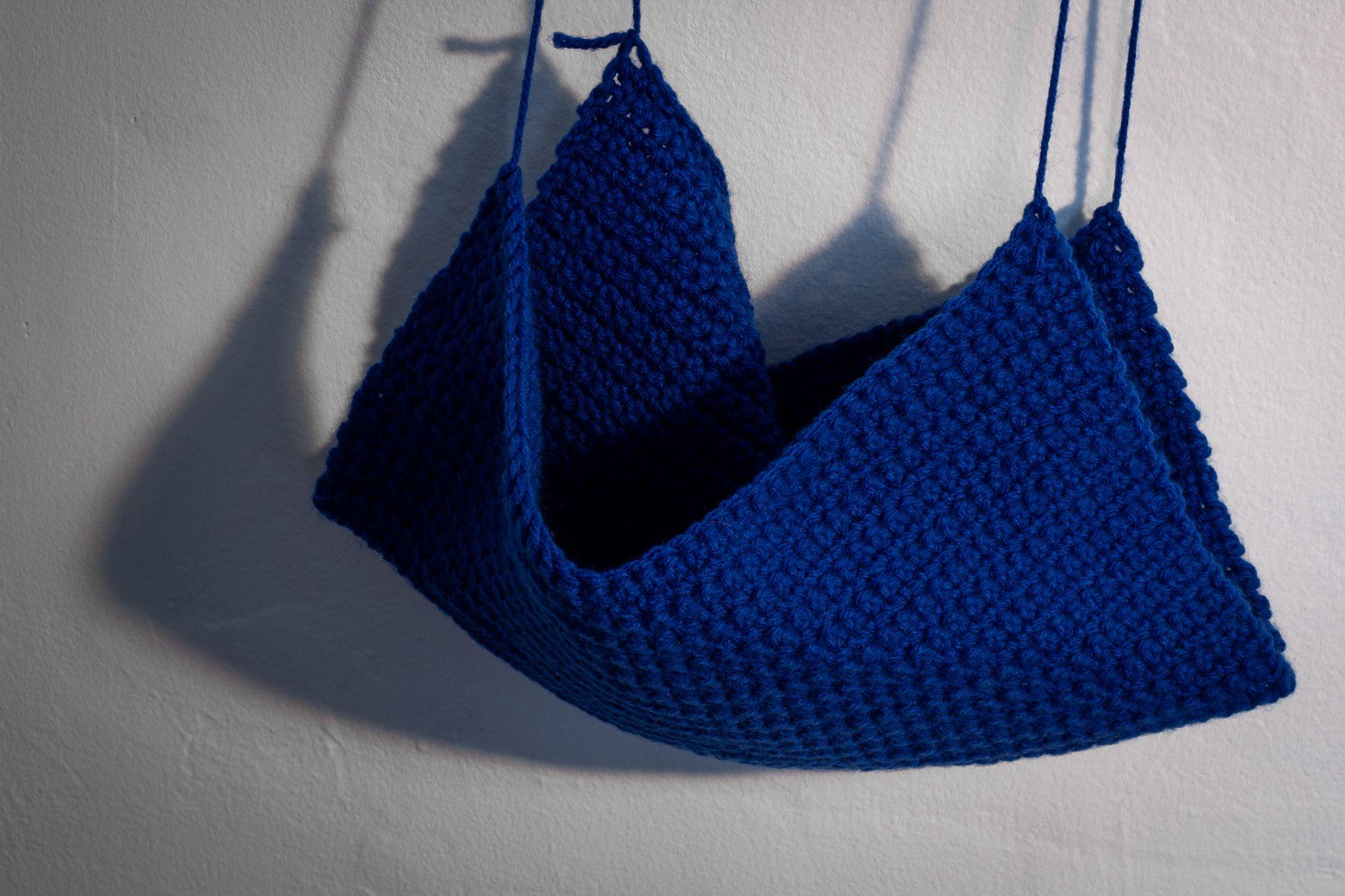 A blue crochet artwork falls from the wall, supported by four threads of yarn, one on each of its corners. 