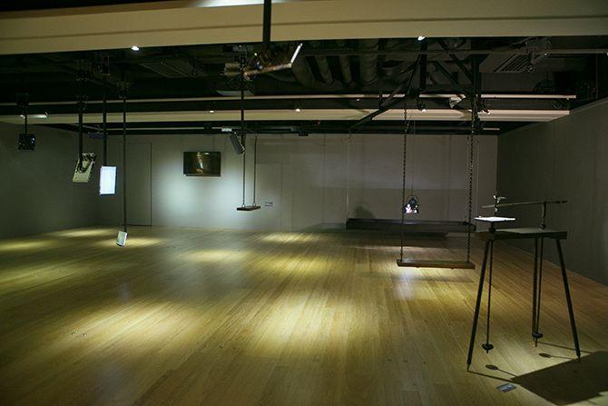 This is a wide shot photo of a room with multiple sparsely located objects in it. On the far left, there is a lighted, white screen. In the middle left, there is a TV turned off and a swing. In the middle right, there is a strip of black material, a stage