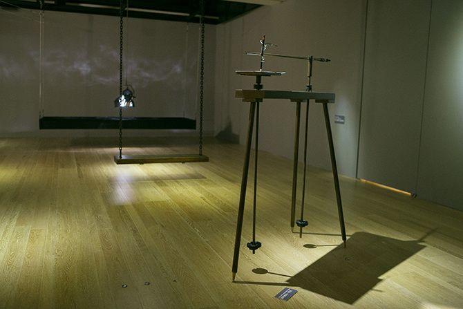 This is a photo of a device with three thin black legs and two long stick-like items that look like ski poles not long enough to touch the ground attached to it. In the background, there is a swing, and behind that, there is a stage light facing downward 