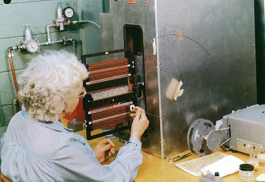 A woman with grey hair sitting in front of a machine feeding copper wire through a frame.