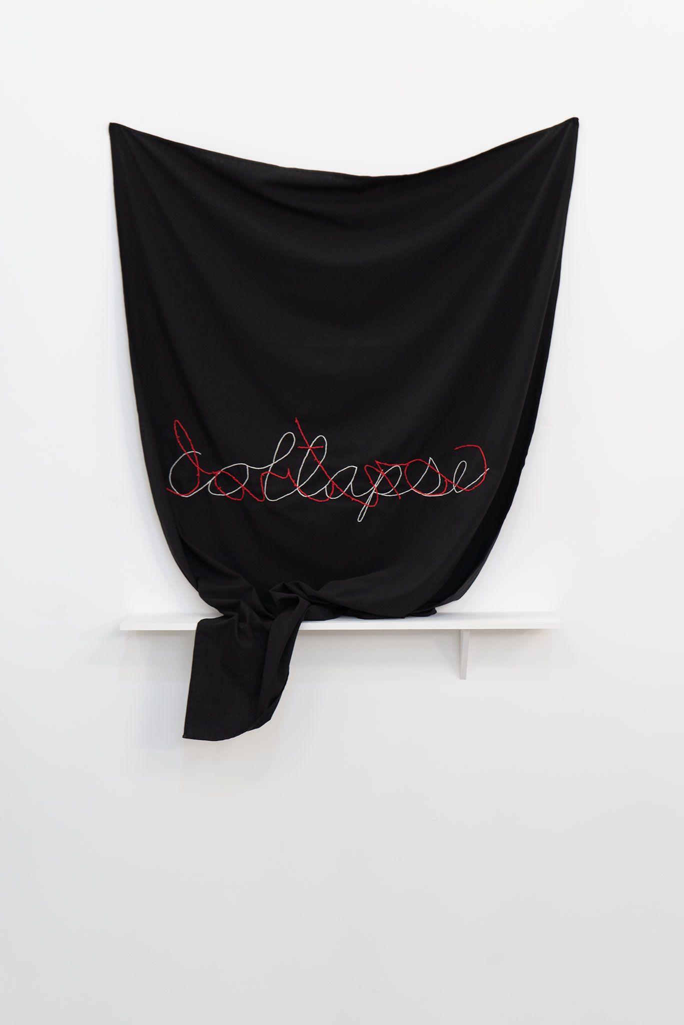 A large piece of black cloth with the word "collapse" emboidered on the front in white. Another word that is hard to make out is embroidered in reverse in red. The fabric if pinned to the wall and draping over a shelf.