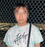 An asian man wearing glasses with medium dark hair. He is wearing a t-shirt that says joker and standing in front of a chainlink fence.