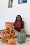 Image of artist in her studio. In front of her is a tall terra cotta ceramic sculpture with broad leaves and coils. Behind her are close up images of water printed and hung on a white wall